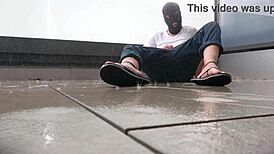 Masked figure masturbates outdoors in wet jeans on a rainy balcony.